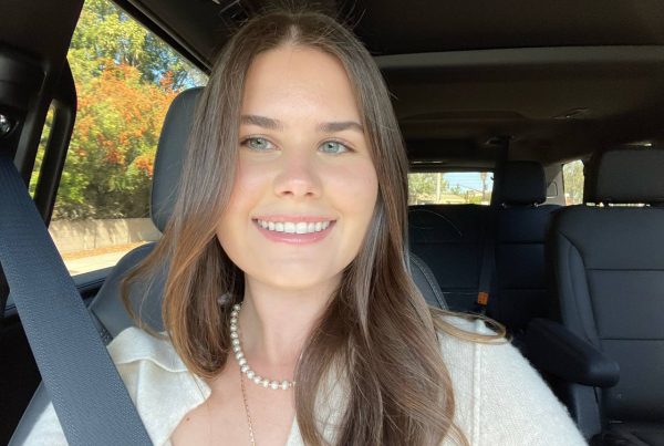 Smiling woman with long, light brown hair and soft highlights, wearing a white top and pearl necklace in a car.