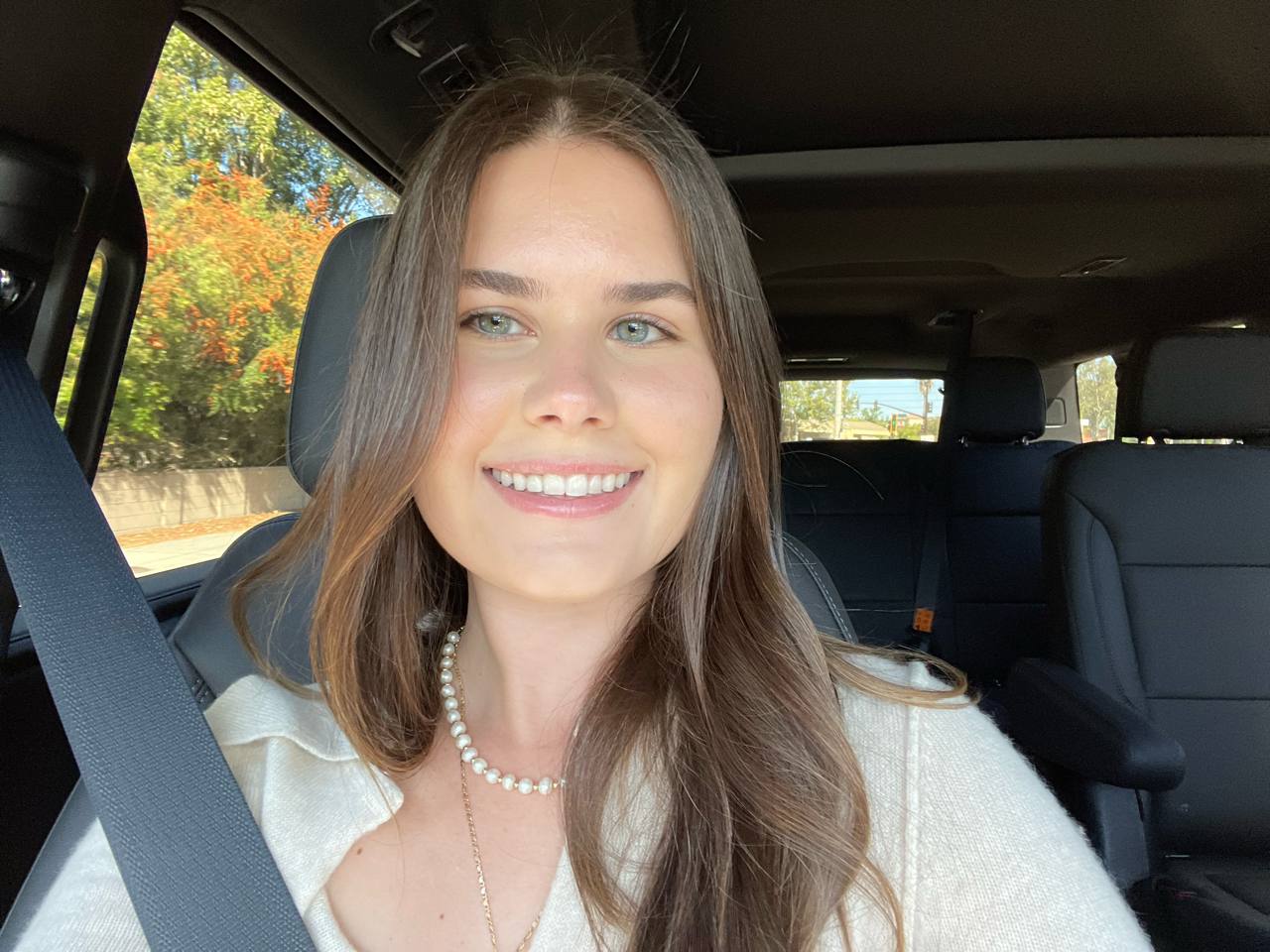 Smiling woman with long, light brown hair and soft highlights, wearing a white top and pearl necklace in a car.
