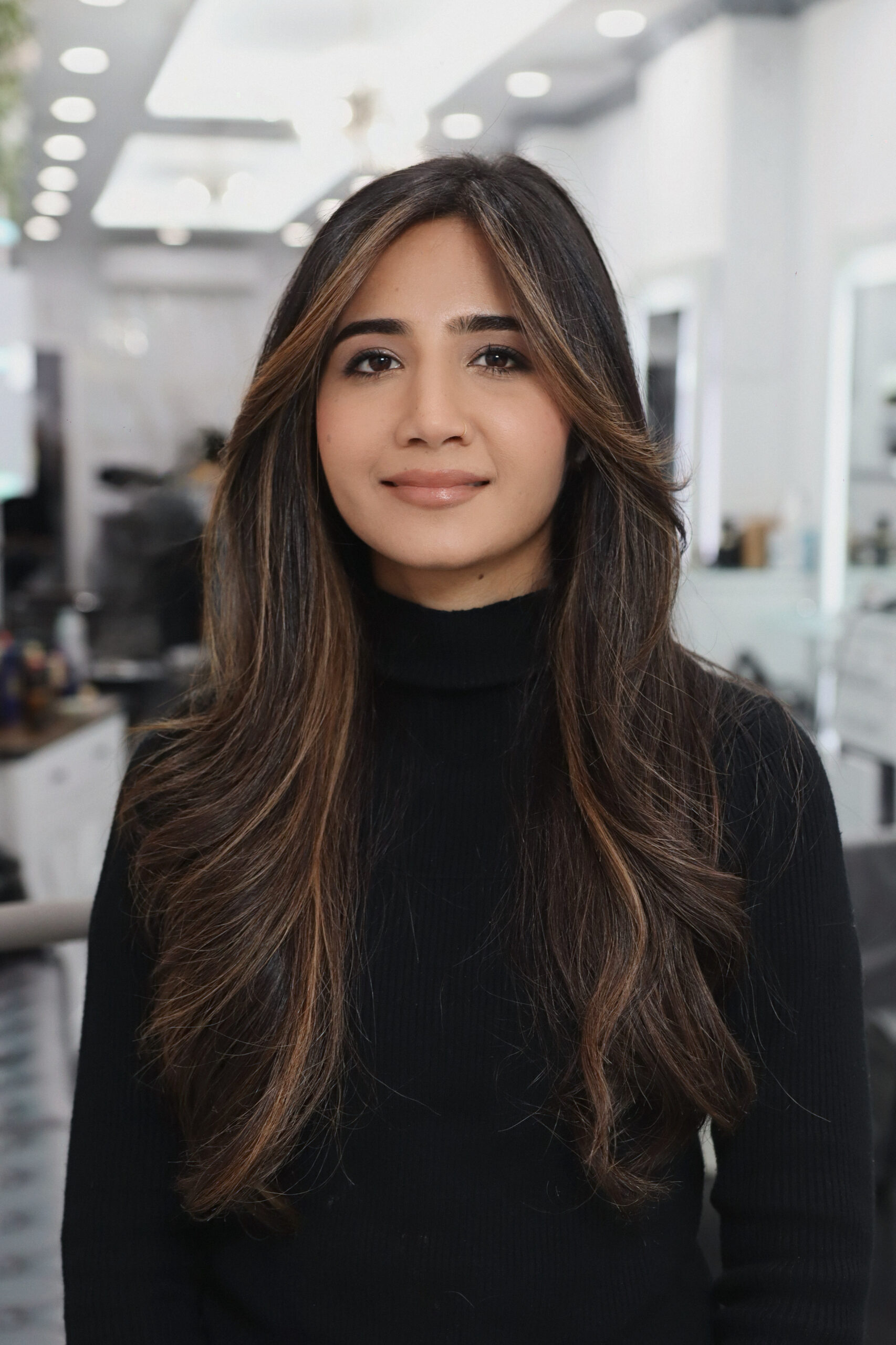 Smiling woman with long, layered brown hair and soft caramel highlights, wearing a black turtleneck inside Davida Salon in NYC.